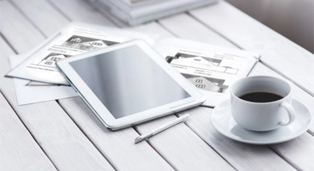 A white tabletop with a set of house plans, an ipad and stylus, and a cup of coffee